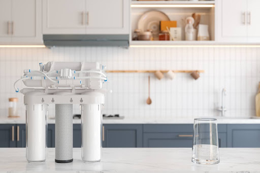 Close-up view of reverse osmosis water filtration system and glass of water on kitchen counter with blurred background.