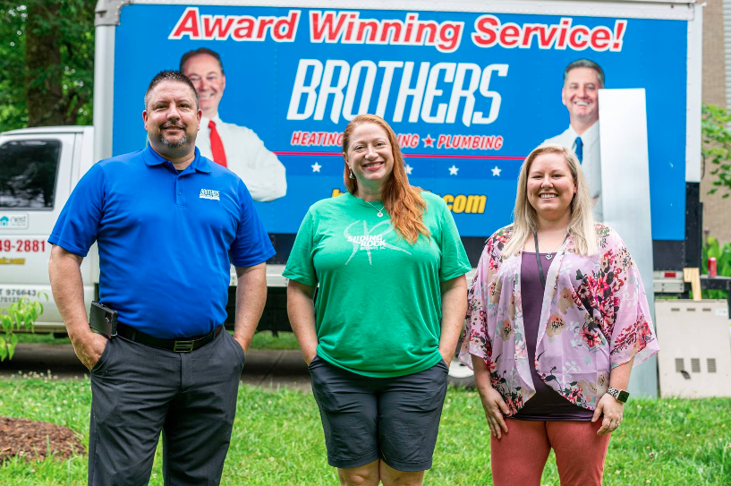 Three people stand in front of a "Brothers Heating & Plumbing" service truck, smiling.